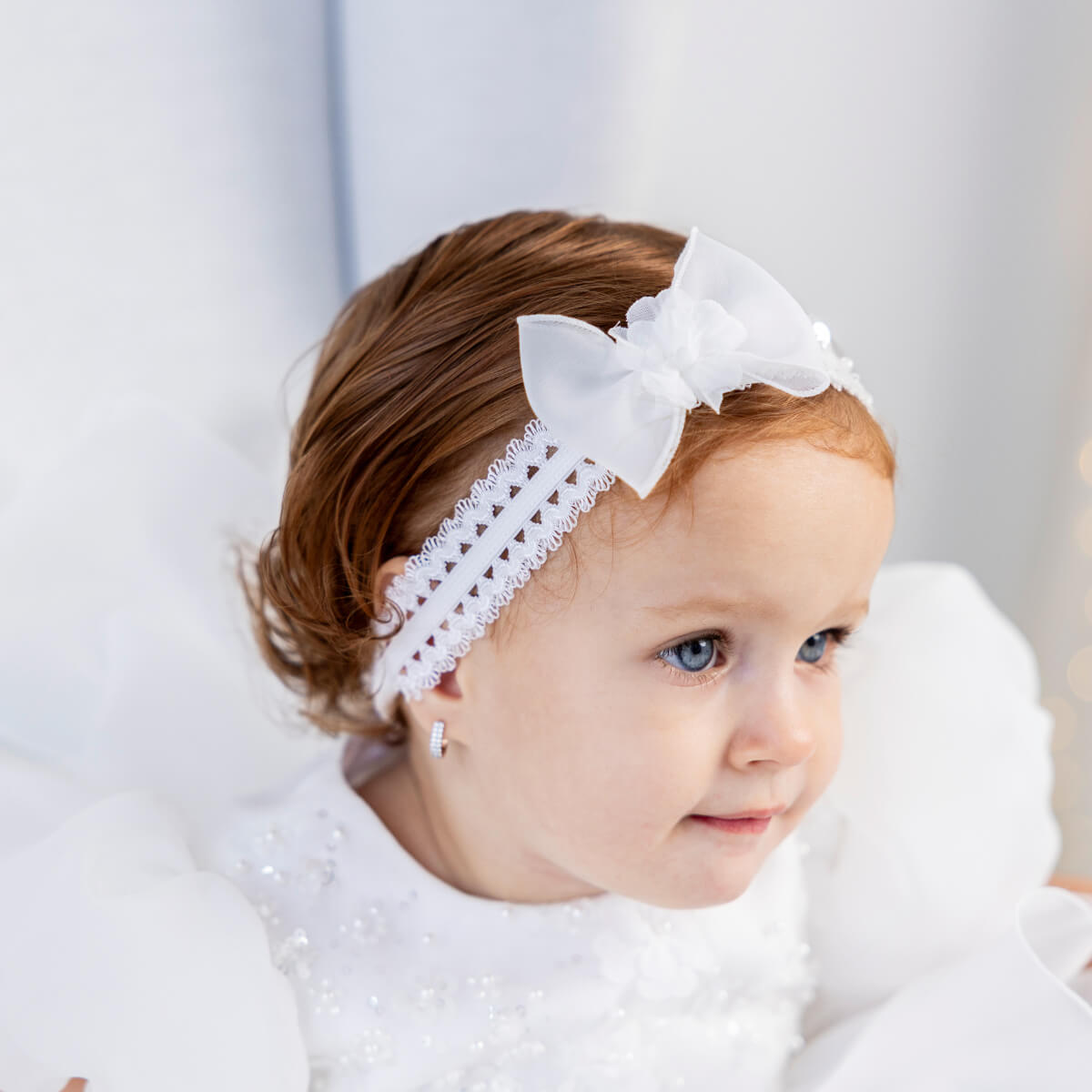 Young girl wearing a bbay headband with a bow, sitting on a white surface.
