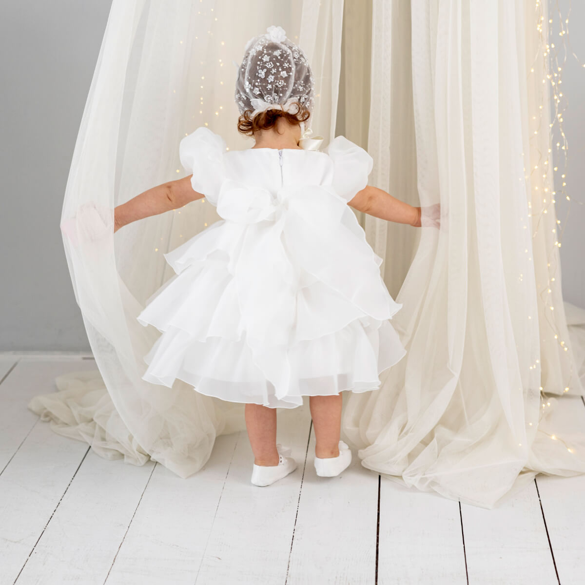 Child in a Baptism Dress with Detachable Train standing between white curtains with fairy lights.