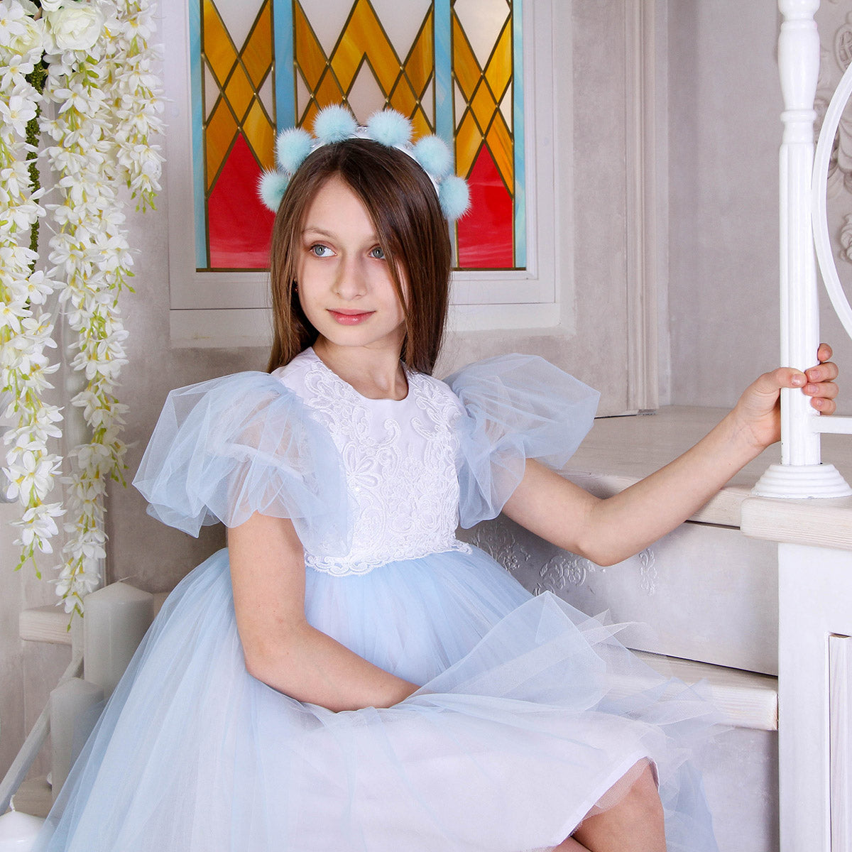 Young girl in a light blue dress with puffed sleeves and a white top, sitting indoors.
