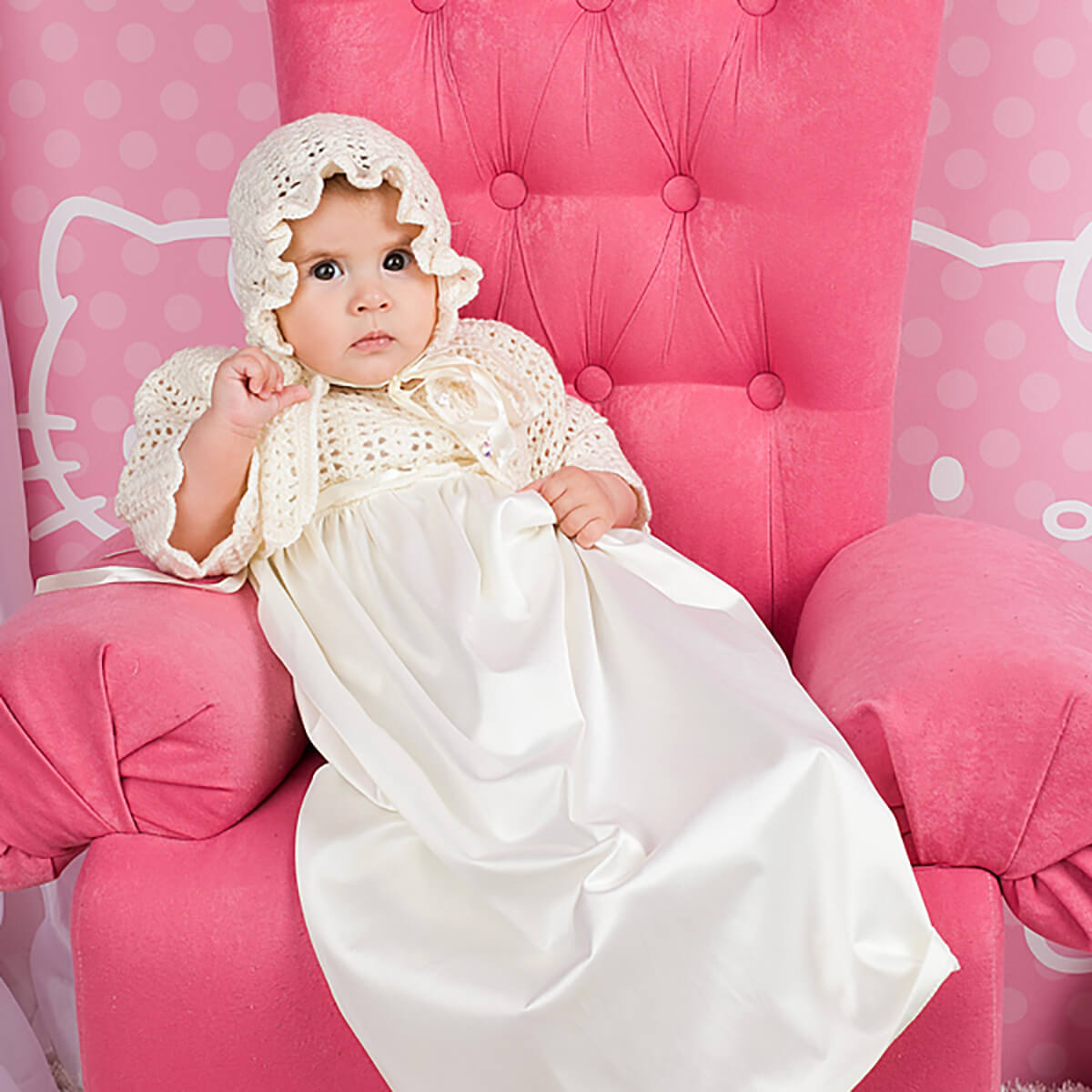 Baby in a Christening Gown for Girls and bonnet sitting on a pink chair with a polka dot background