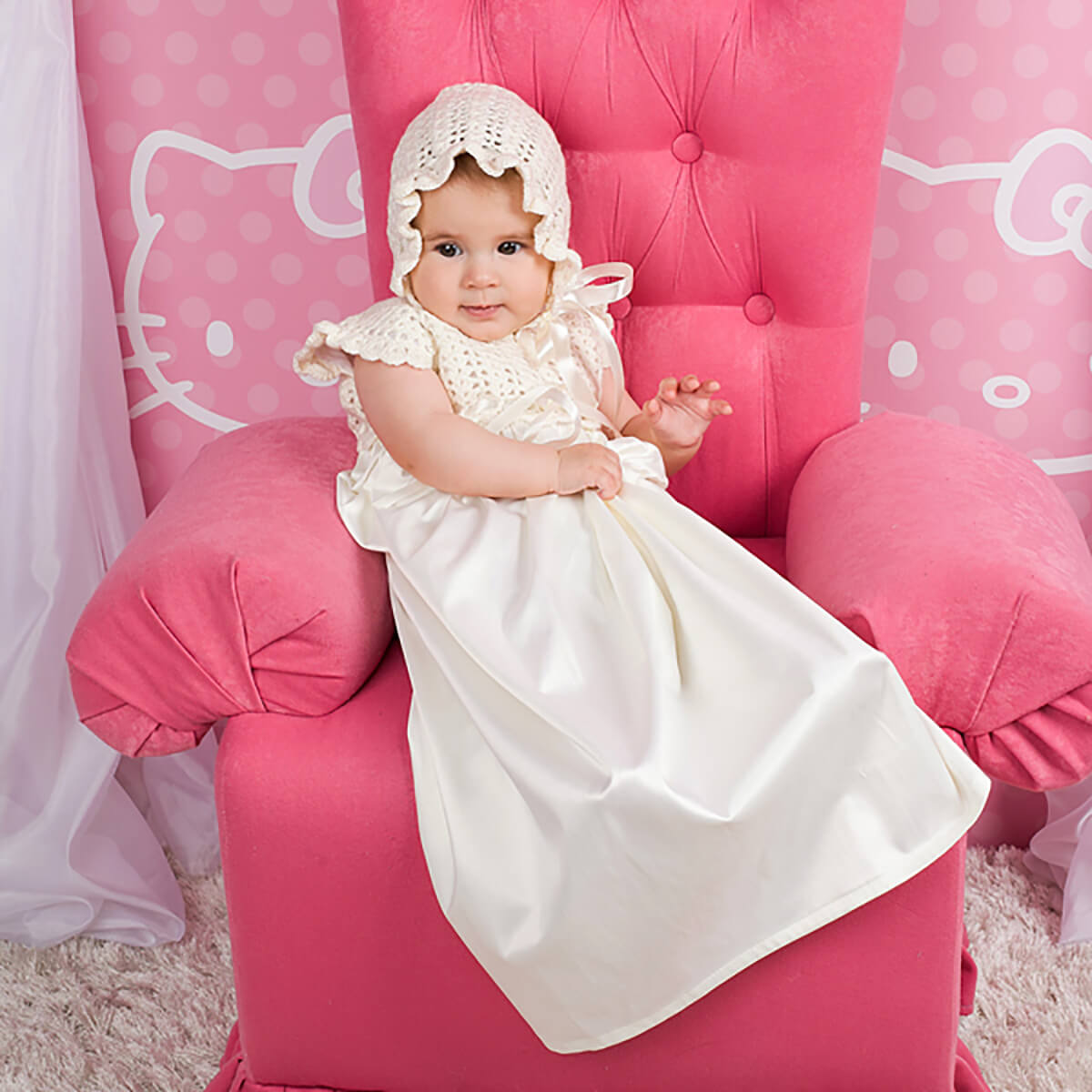 Baby in a Christening Gown for Girls and bonnet sitting on a pink chair with Hello Kitty design.