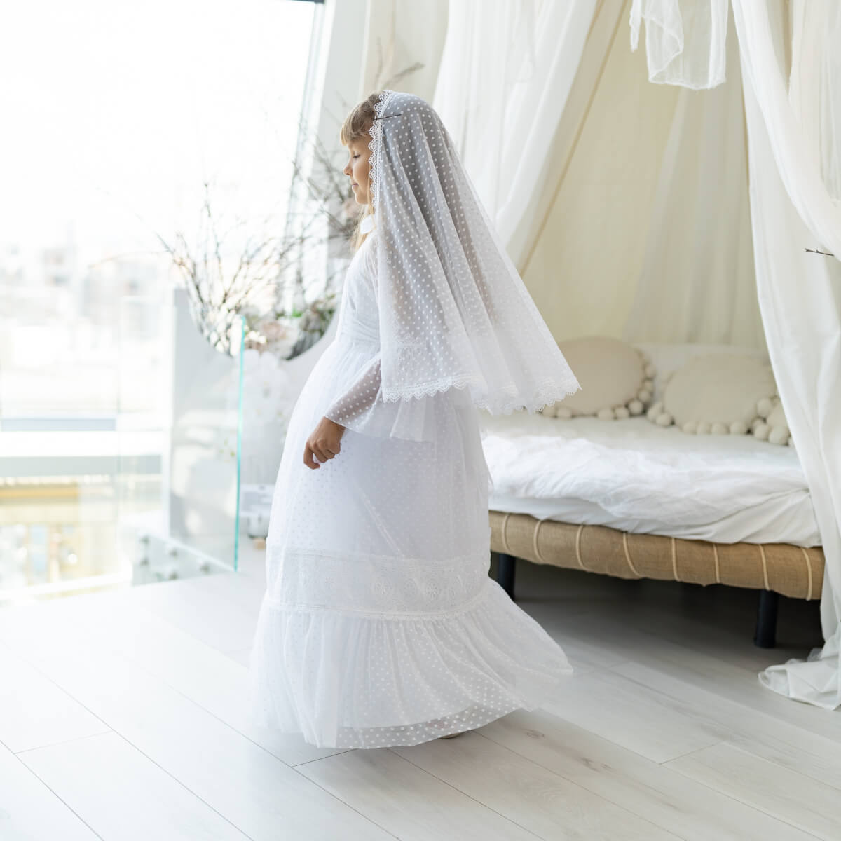 Woman in a white dress with a First Communion Mantilla Veil standing in a bright room with large windows.