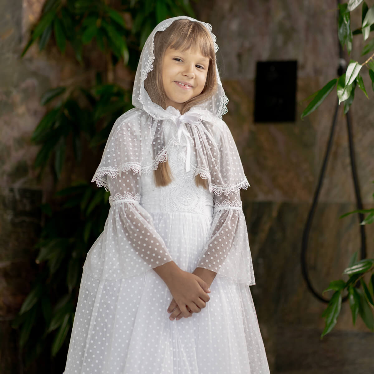 Young girl in a white dress with a First Communion Mantilla Veil standing against a stone wall.