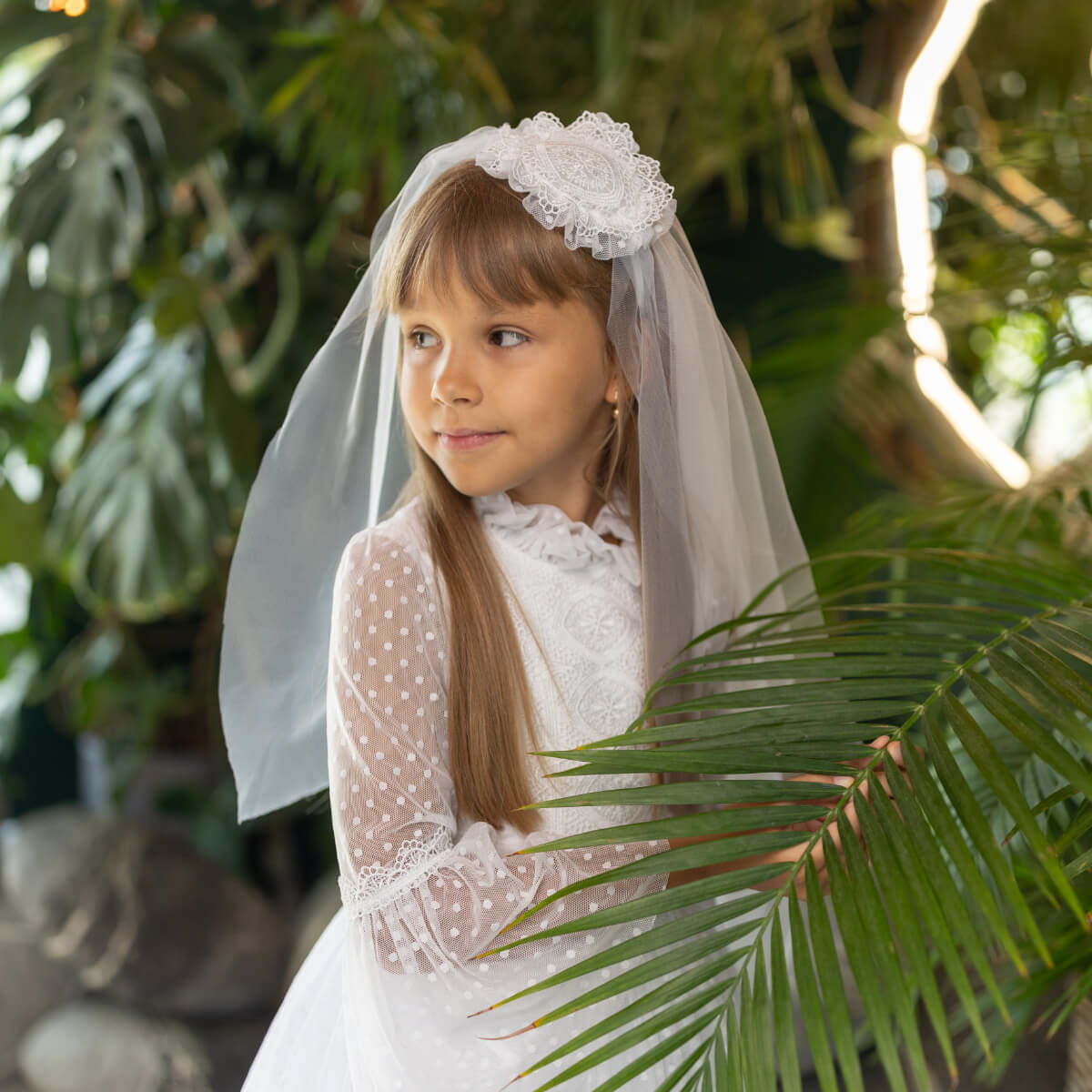 Young girl in a white dress with a First Communion Mantilla standing among green plants.