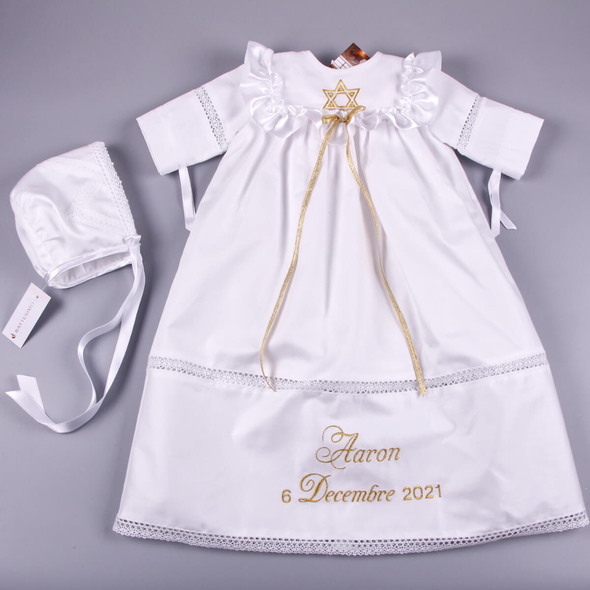 White baby dress with gold embroidery and matching bonnet on a gray background