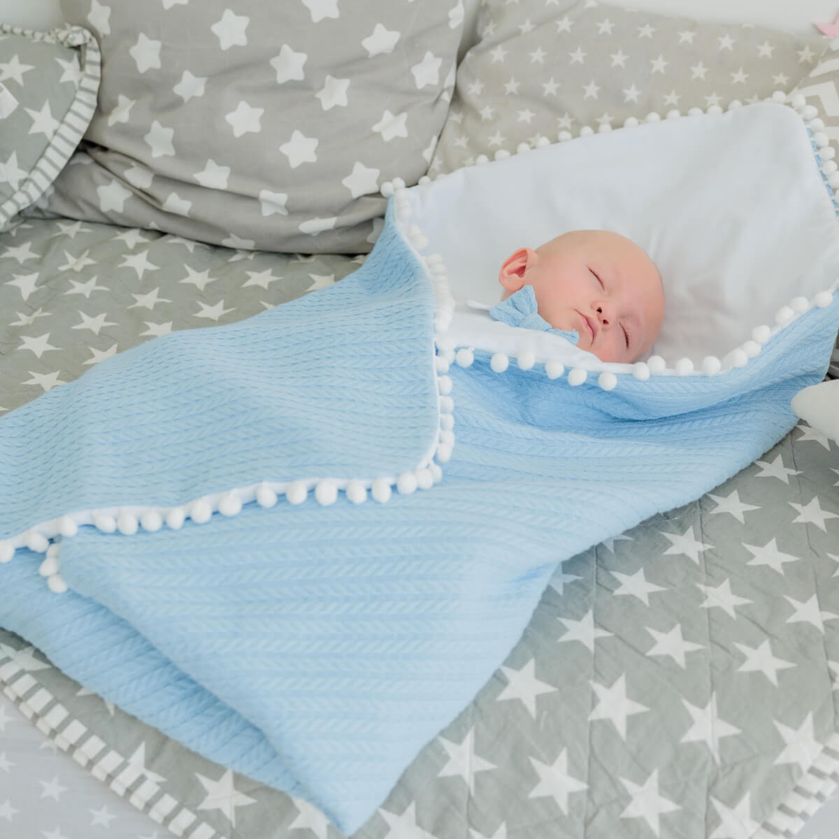 Newborn baby sleeping under a Coming Home Blanket with white pom-poms on a gray blanket with white star patterns.