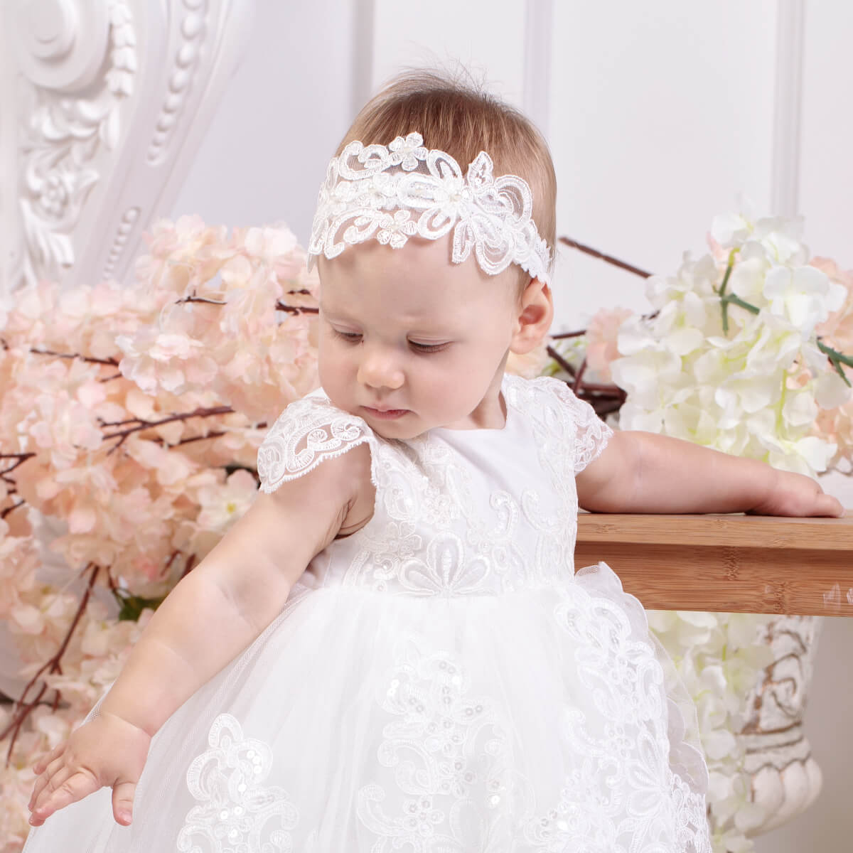 Baby in a white lace dress and baby headband with lace and pearls sitting on a floral background