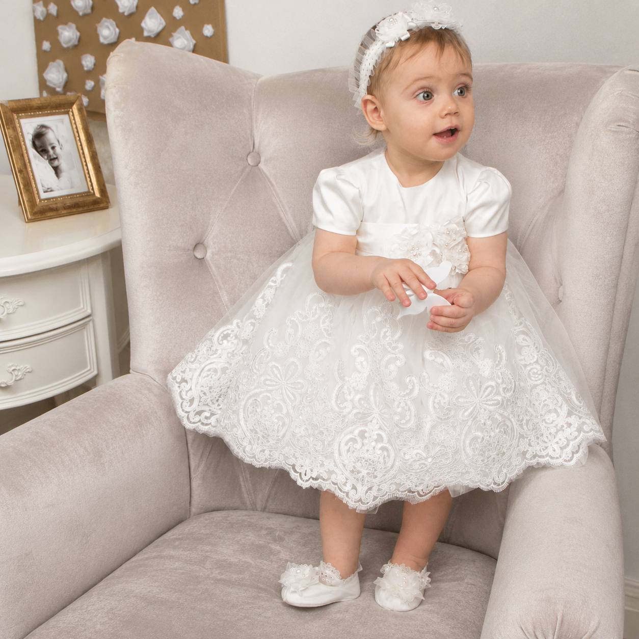 Baby in a white lace dress sitting on a gray armchair.