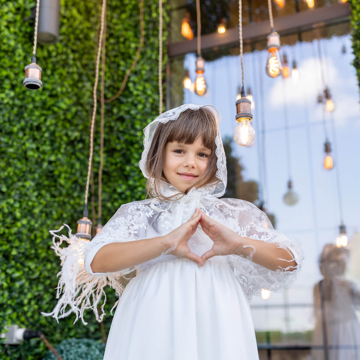 Young girl in a white dress with a Holy Communion Mantilla, standing in an outdoor setting with greenery and hanging lights.