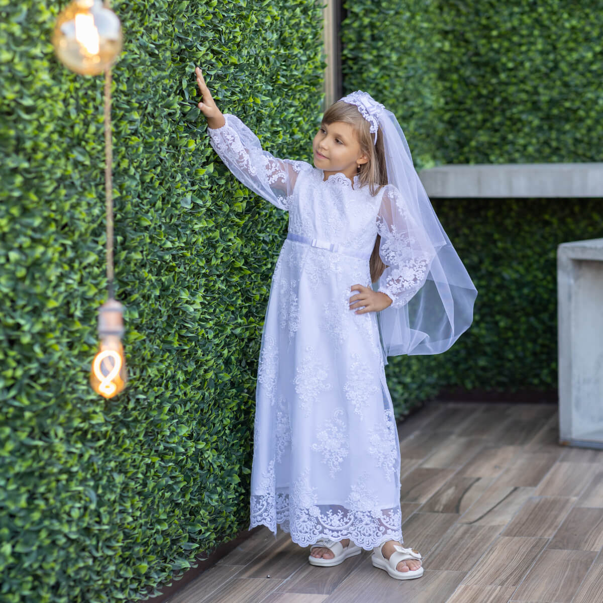 Young girl in a white lace Simple Lace Communion Dress standing outdoors against a green hedge.