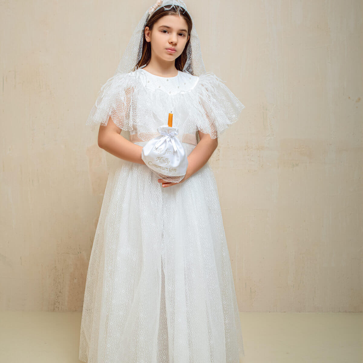 Young girl in a white dress with a First Communion Veil with Bow holding a small white bag against a beige background