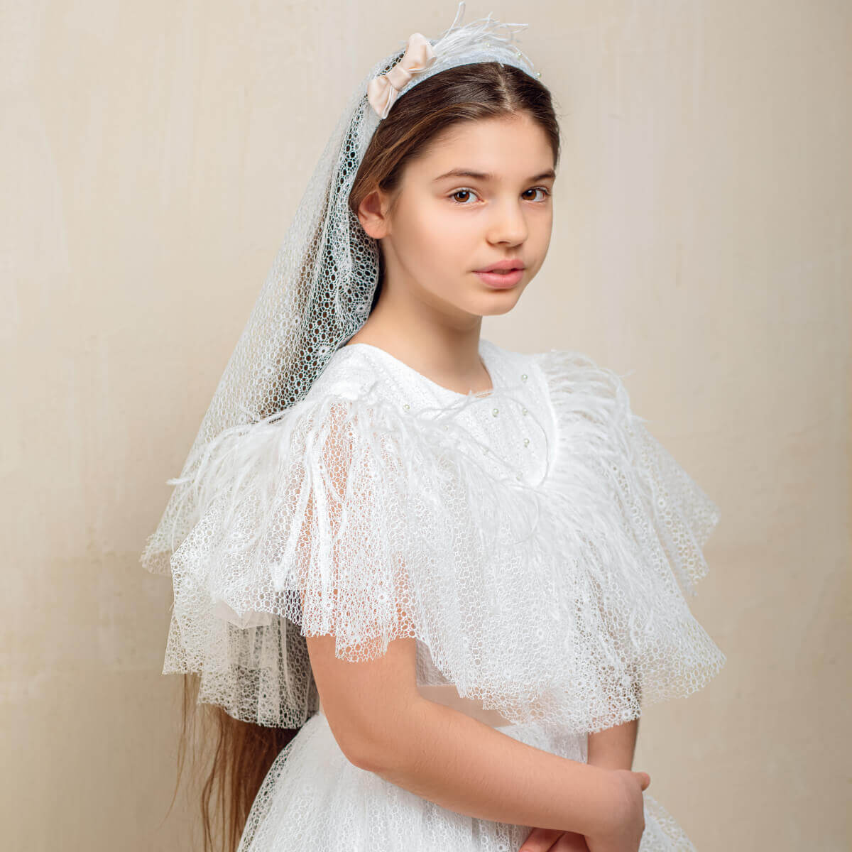 Young girl wearing a white lace dress with a matching First Communion Veil with Bow against a beige background