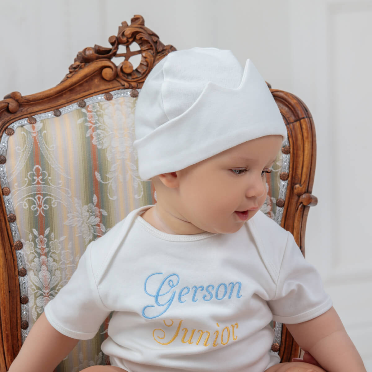Baby wearing a white hat and a Coming Home Bodysui with embroidered text, sitting on a wooden chair.
