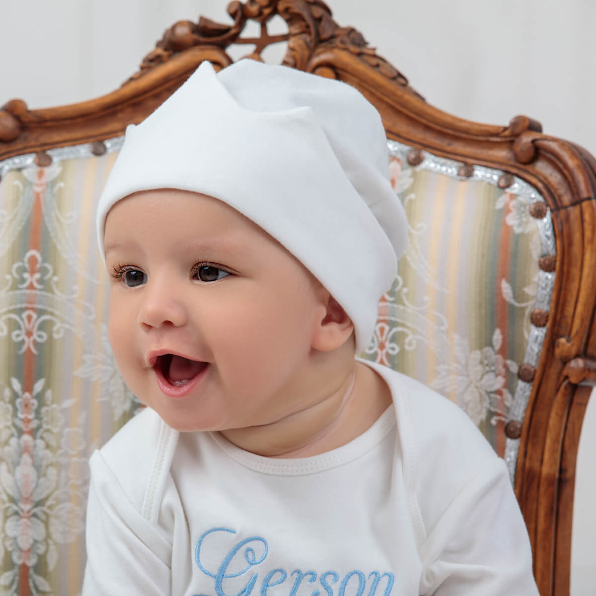 Baby wearing a white hat and Coming Home Bodysui with 'Gerson' on it, sitting on a decorative chair.
