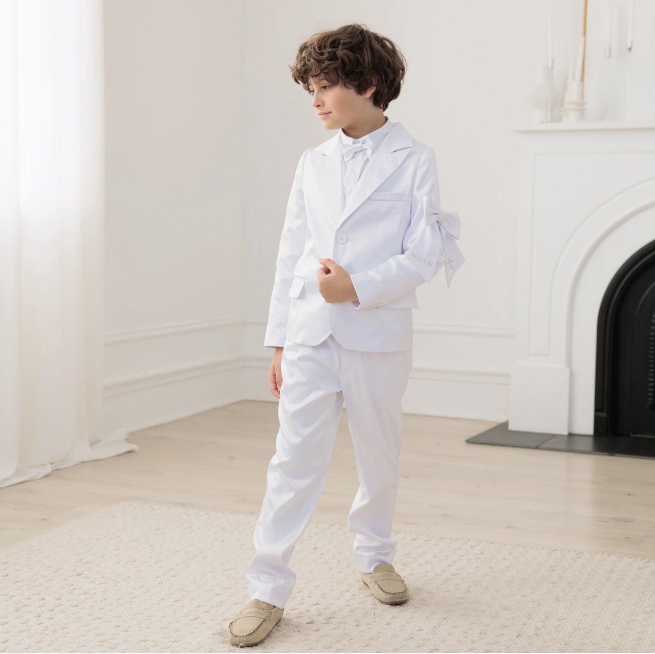 Child in a white communion suit standing in a bright room with a fireplace.