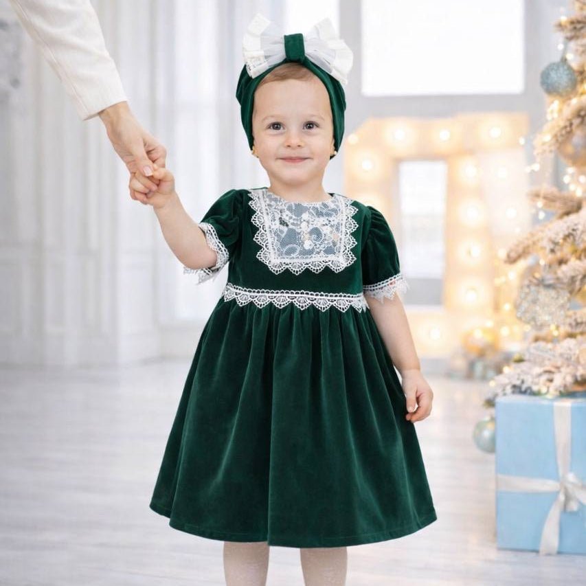 Child in a green dress with white lace details standing in a festive indoor setting.