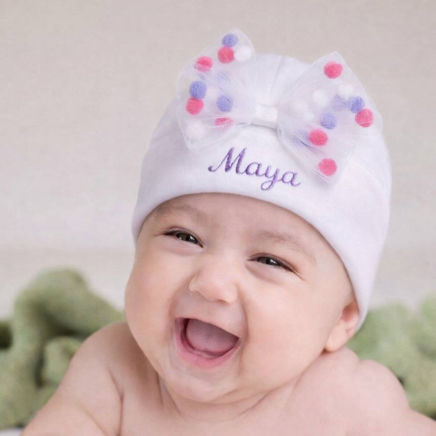 Baby wearing a white hat with colorful pom-poms and the name 'Maya' on a light background