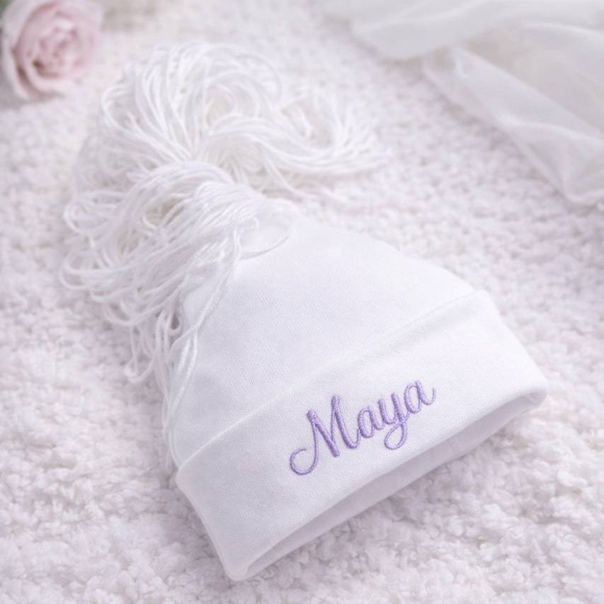White beanie with 'Maya' embroidered on it, placed on a fluffy white surface.