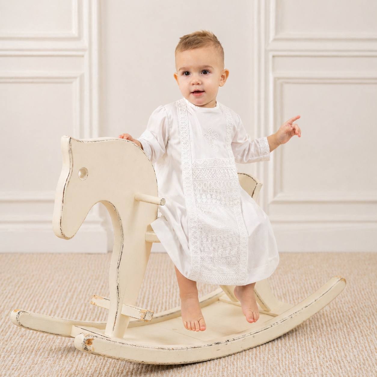 Baby in a white boy baptism outfit sitting on a wooden rocking horse against a white wall.