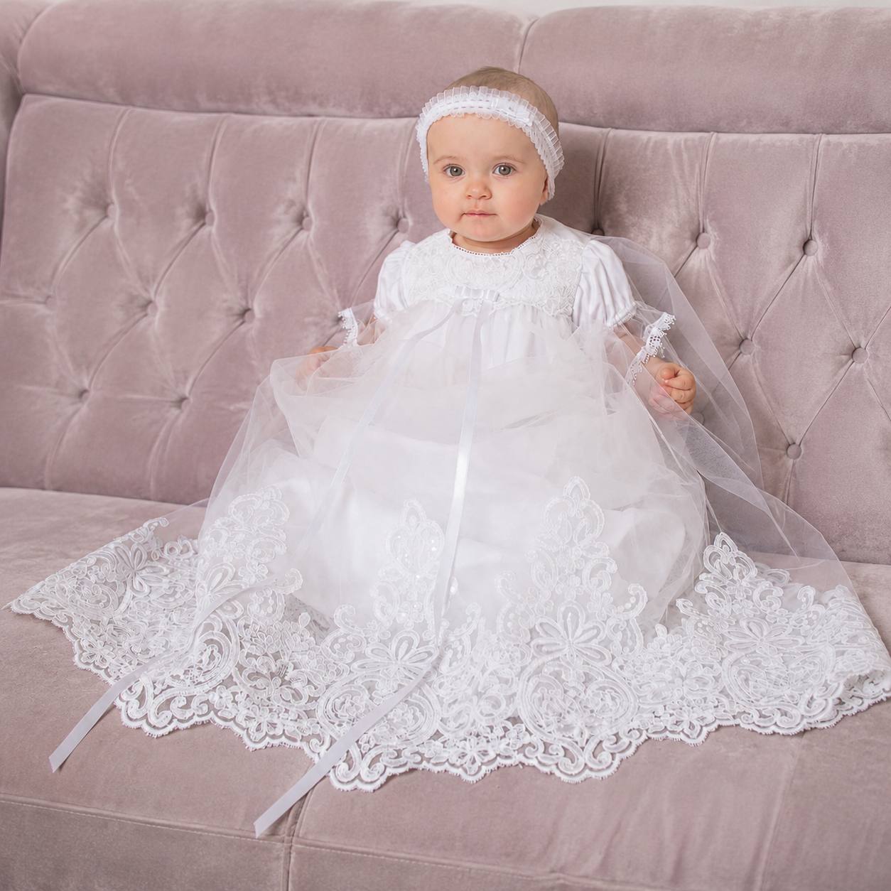 Baby in a white lace baptism gown. dress sitting on a pink couch