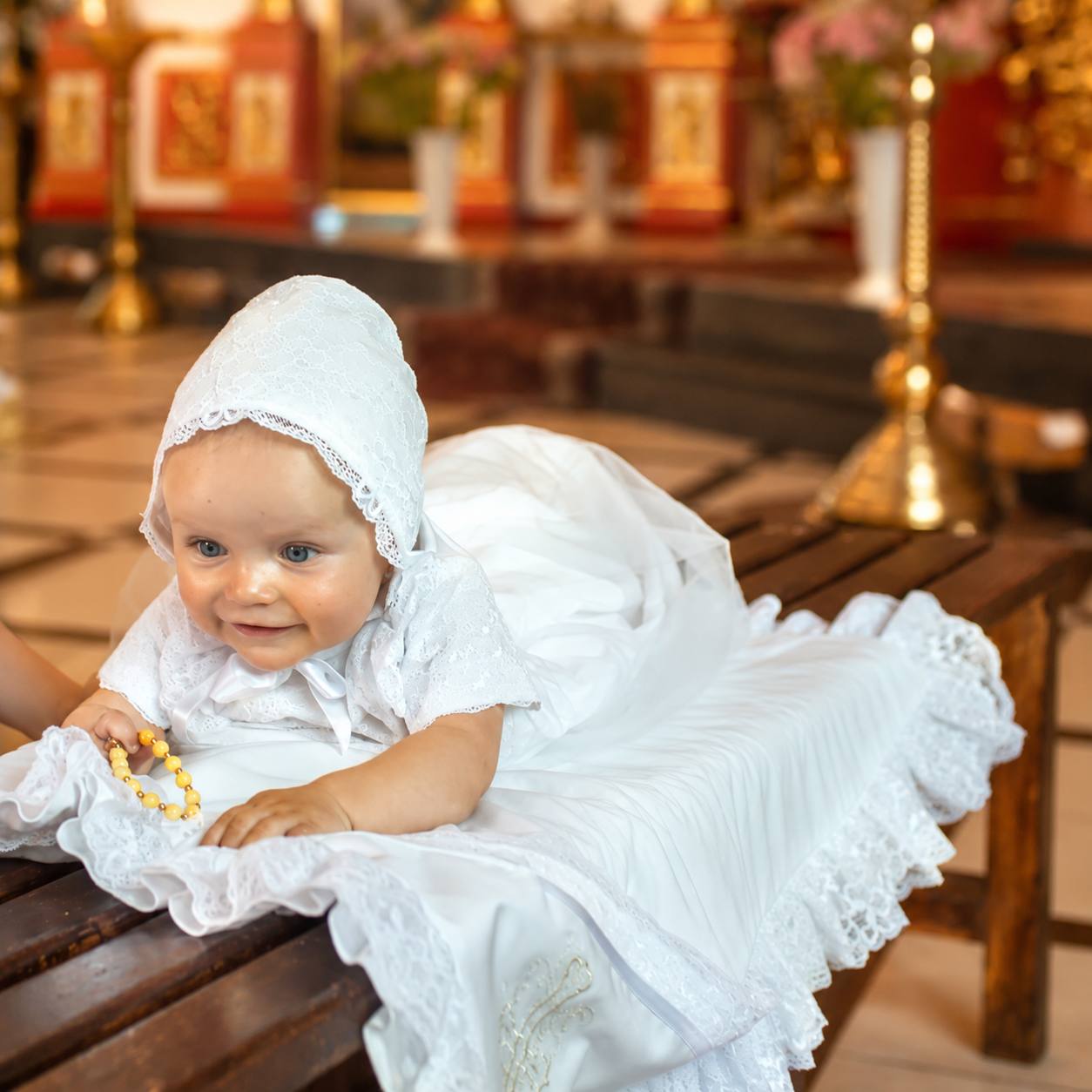 Baby in a white dress and headscarf sitting on a wooden bench in a decorated room.