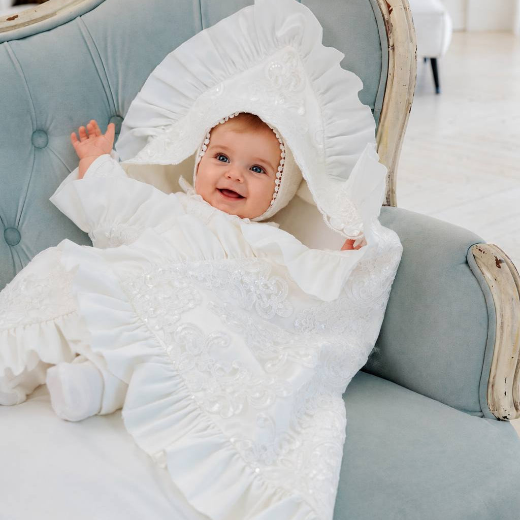 A baby wearing a white embroidered bonnet and matching white baby outfit, reclining on a light blue tufted vintage-style armchair.