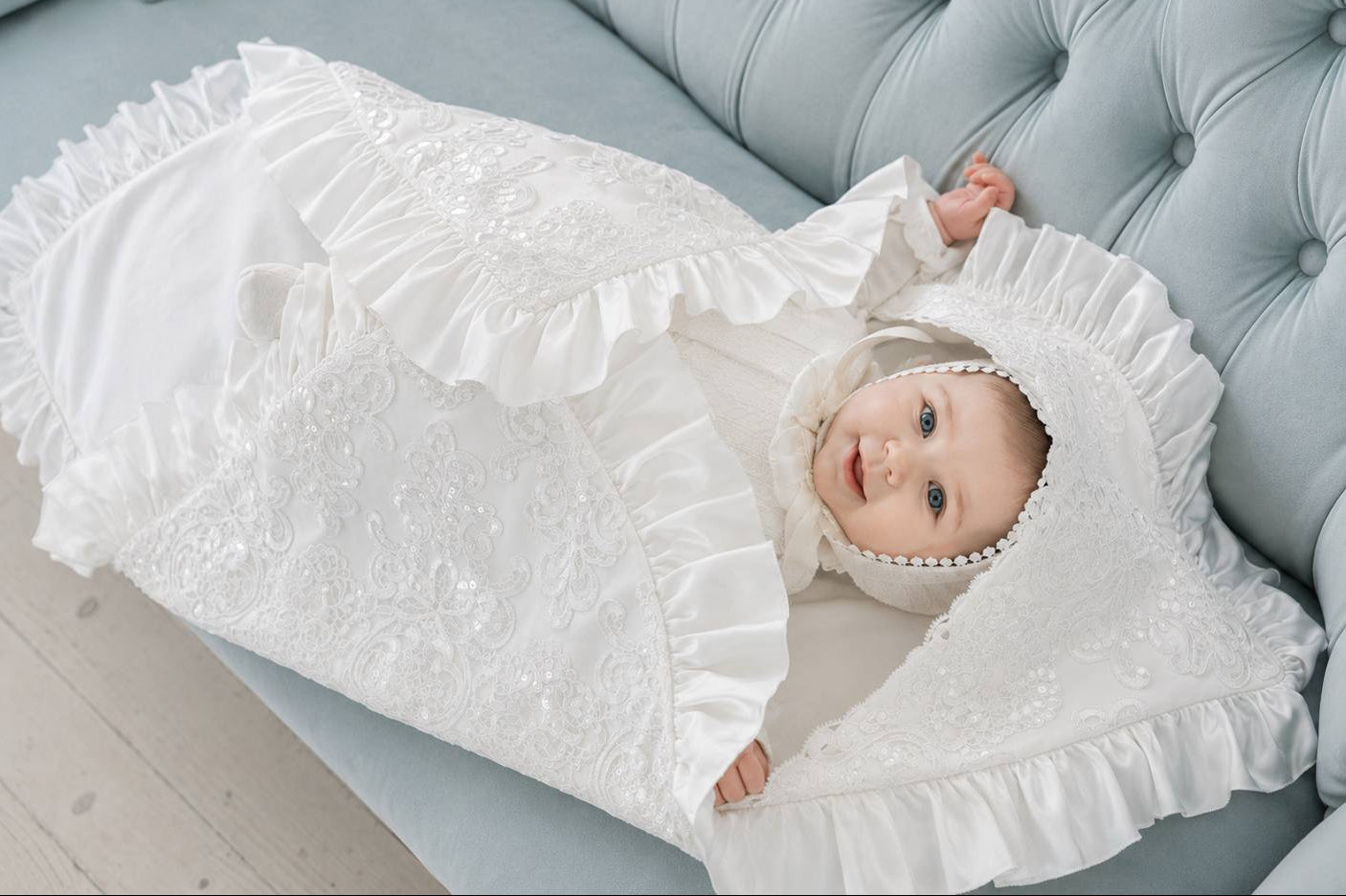 A baby wrapped in a white lace and ruffle christening blan and bonnet, lying on a light blue tufted sofa.