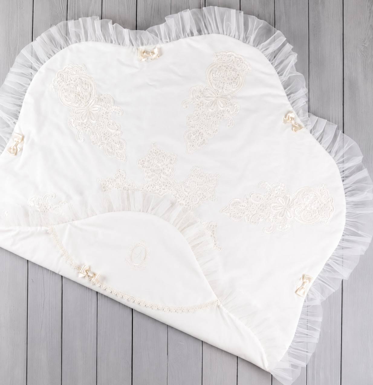 An off-white baby blanket with delicate lace embroidery and a ruffled tulle border, laid flat on a gray wooden plank surface.