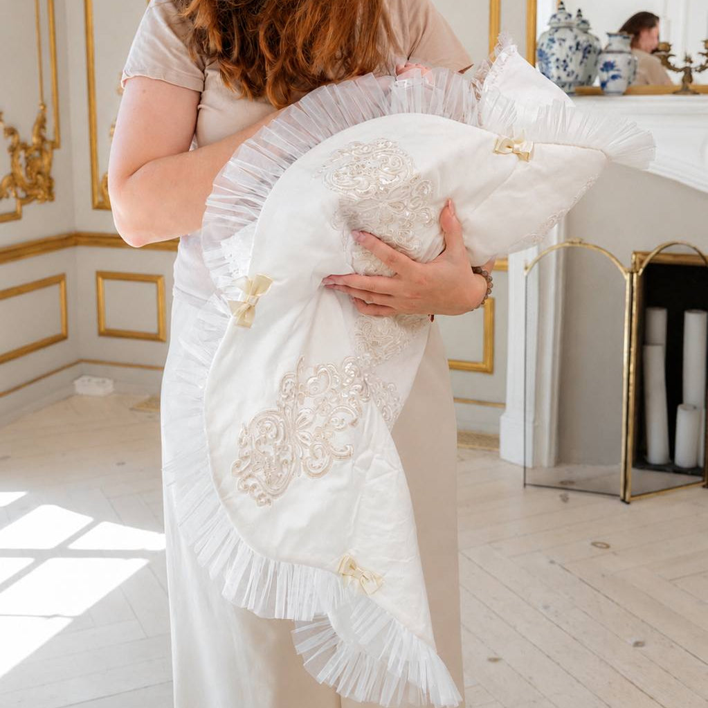A woman in a cream-colored dress holds a white fringed baby blanket with gold embroidered floral patterns in a bright, elegant room with gold-trimmed walls.