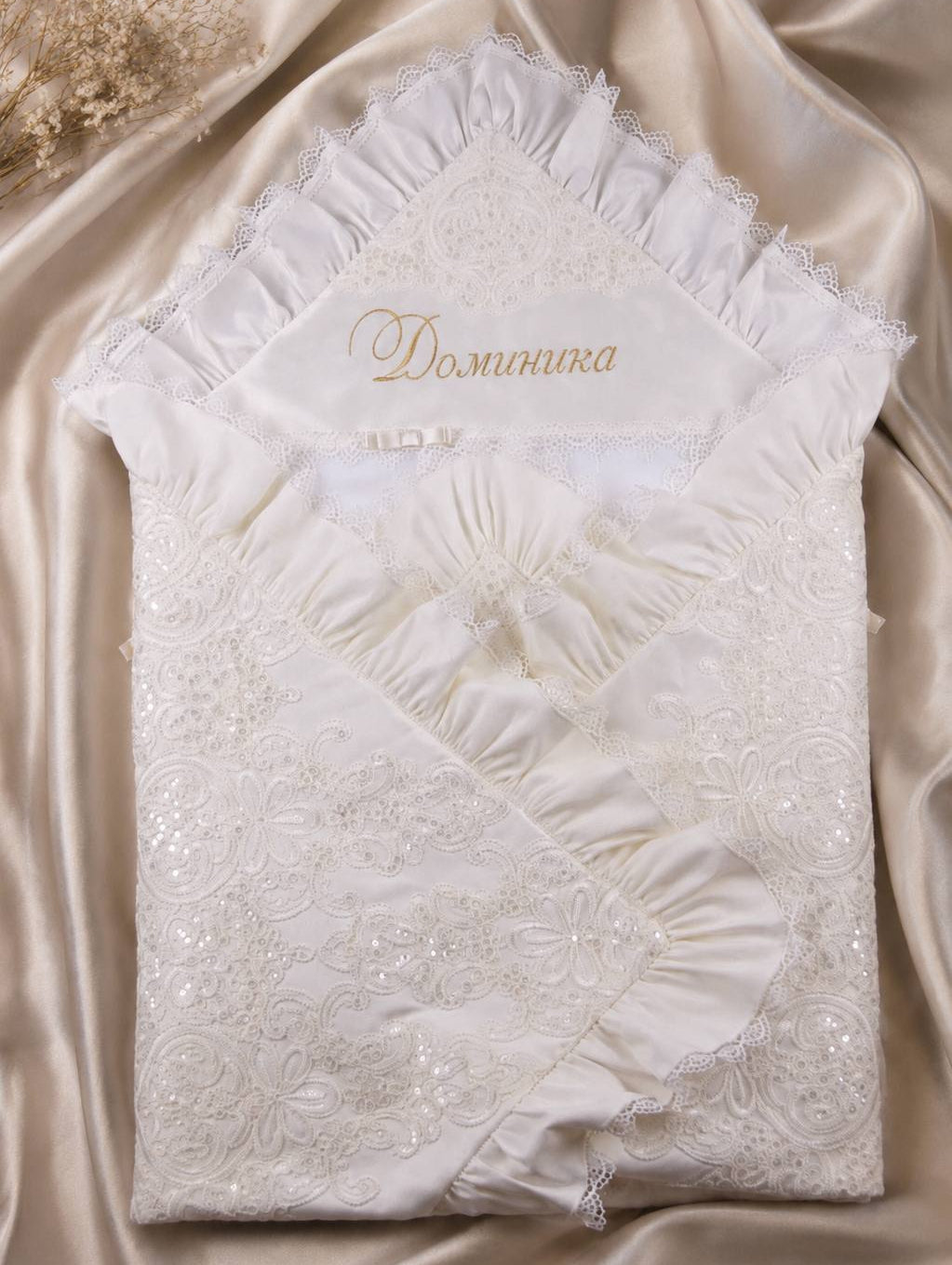 An off-white embroidered lace baby blanket with ruffled edges and a gold embroidered monogram 'D. O.