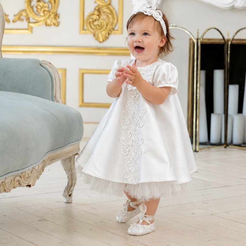 A toddler girl wearing a white satin baptism dress with lace embroidery, a matching white headband, and white baby shoes, standing in a luxurious room with gold-trimmed walls and a fireplace.