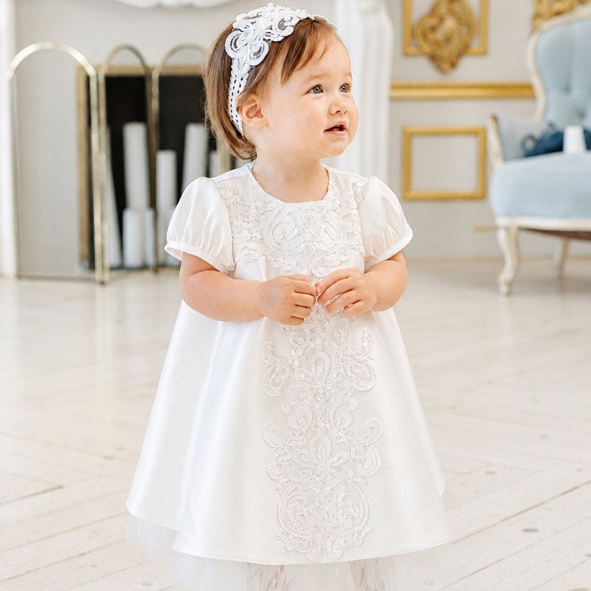 A toddler girl wearing a white lace and satin baptism dress with a matching white headband, standing in a bright, elegant room with a fireplace and gold-trimmed walls.