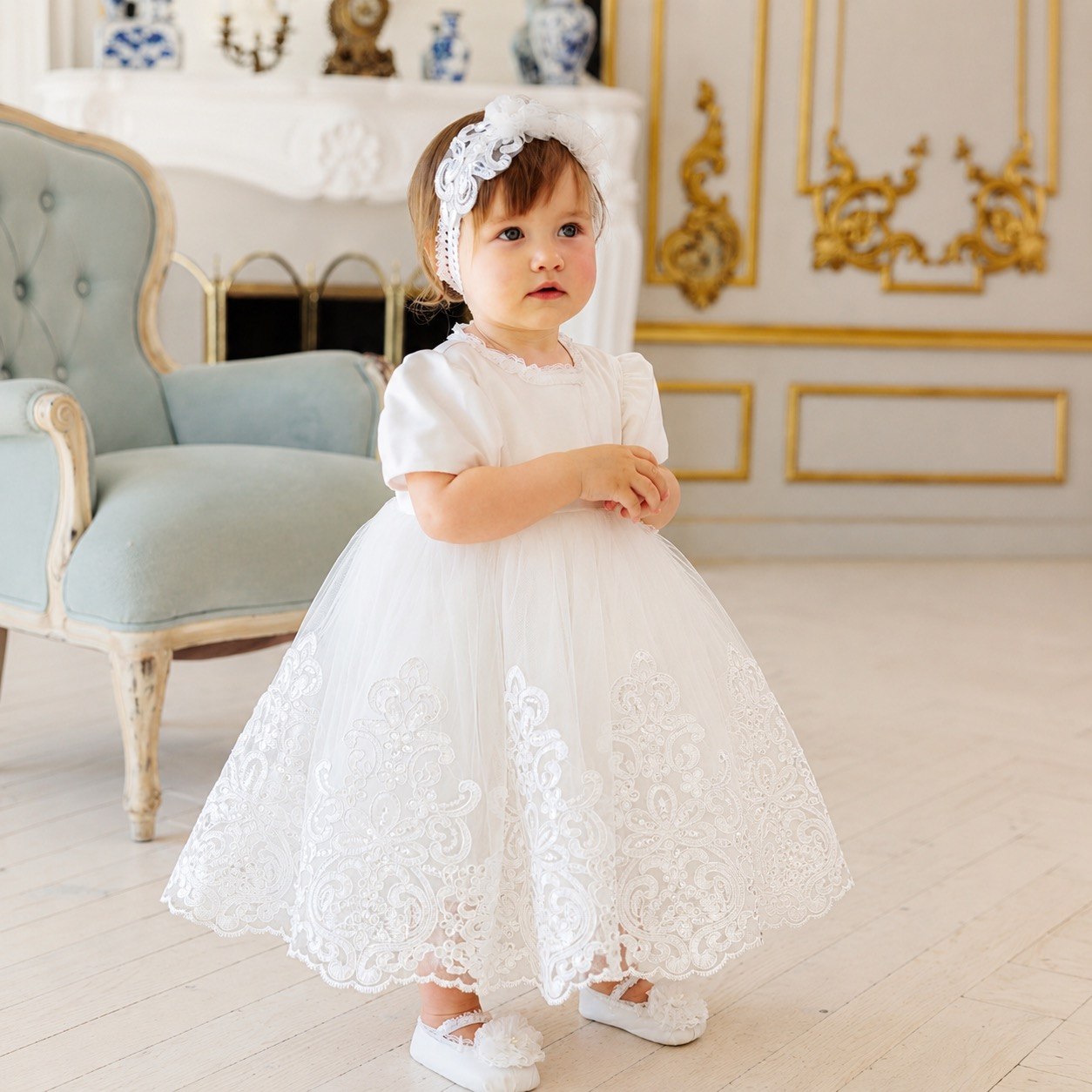 A toddler girl wearing a white short-sleeve lace dress with a matching white floral headband and white shoes