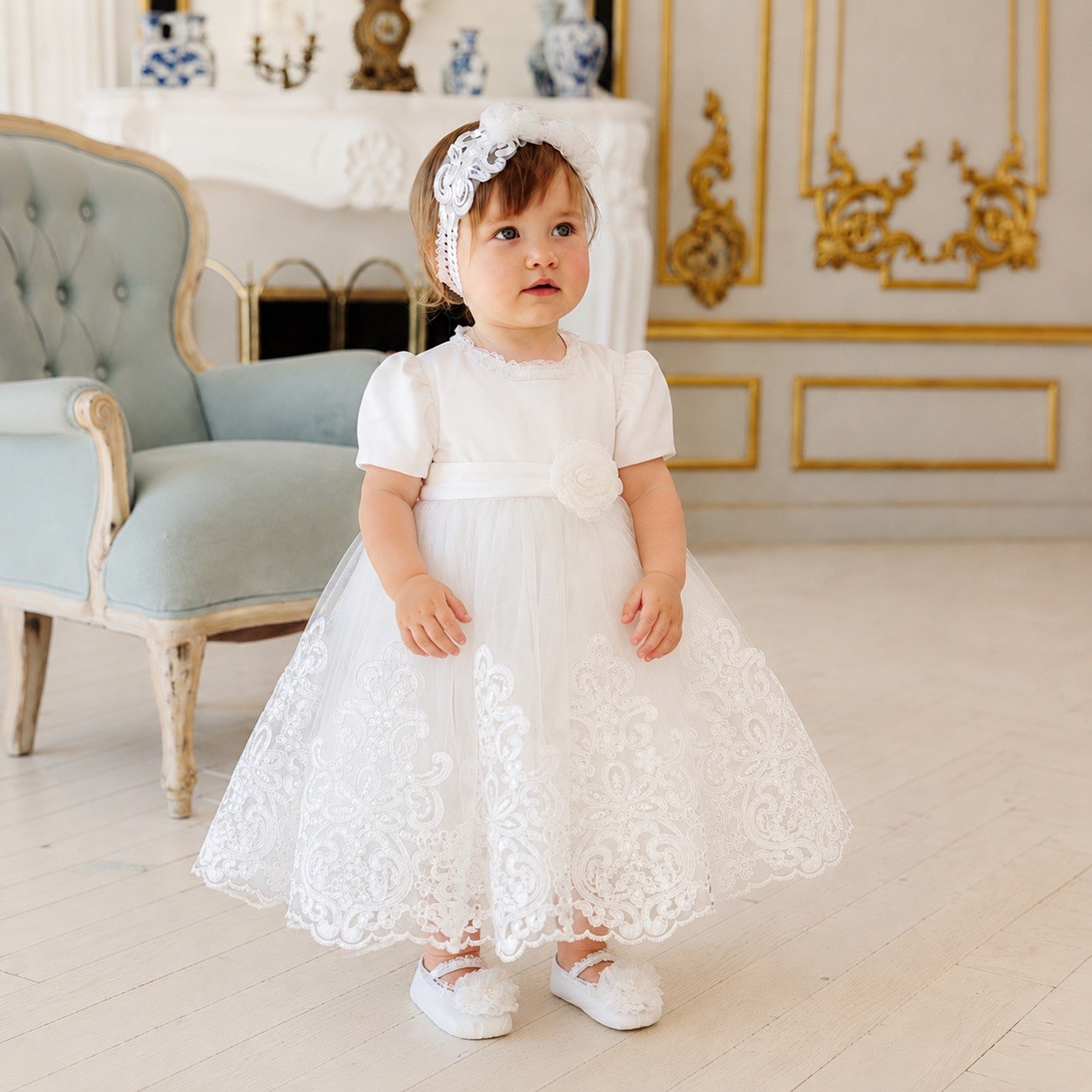 A toddler girl wearing a white lace and tulle first communion dress with a matching white lace headband, standing in a beautifully decorated room with a light blue armchair and gold-trimmed walls.