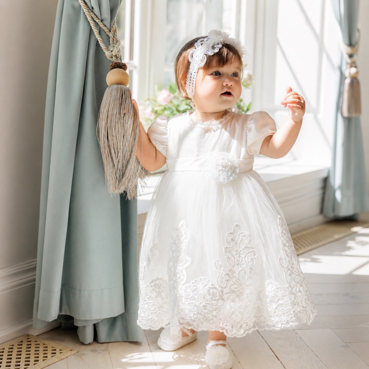 A toddler girl wearing a white lace and tulle first communion dress with a matching white lace headband 