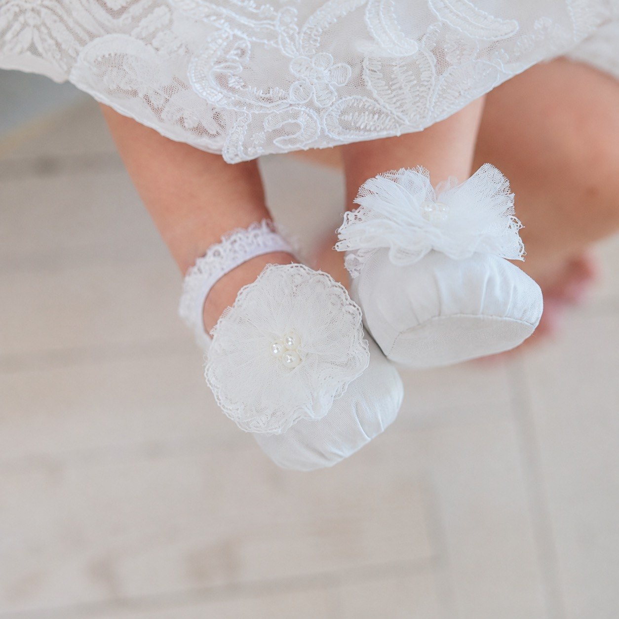 White baby booties with lace trim and ruffled flower details, shown on a child's feet under a white lace dress.
