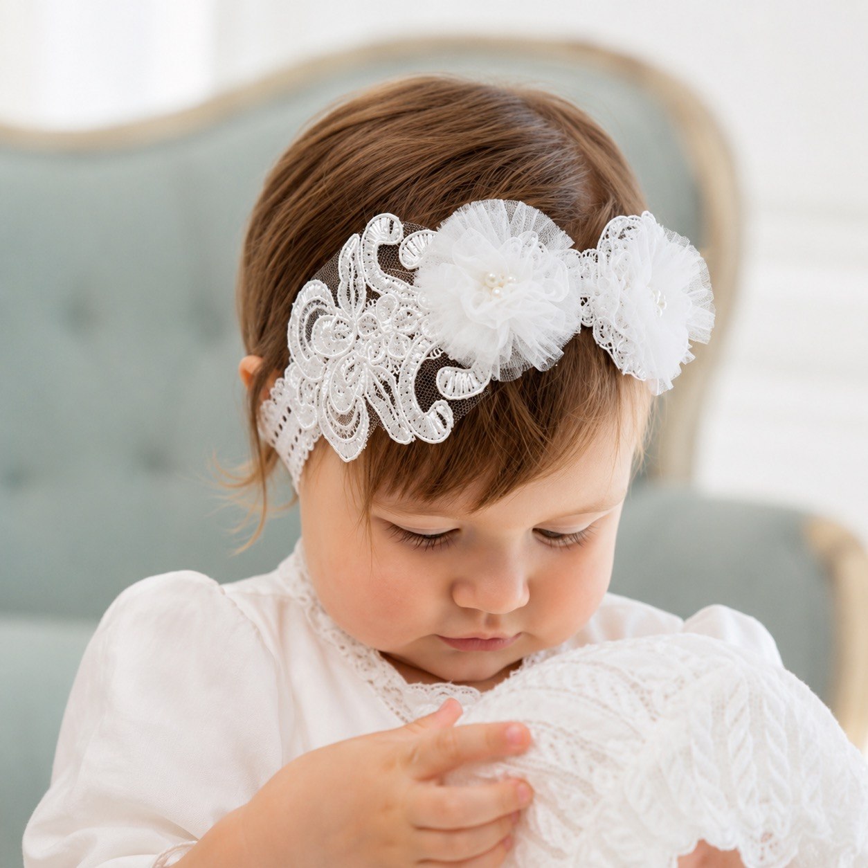 A toddler wearing a white lace headband with two large ruffled flowers, looking down while holding a white lace item in a softly lit indoor setting.