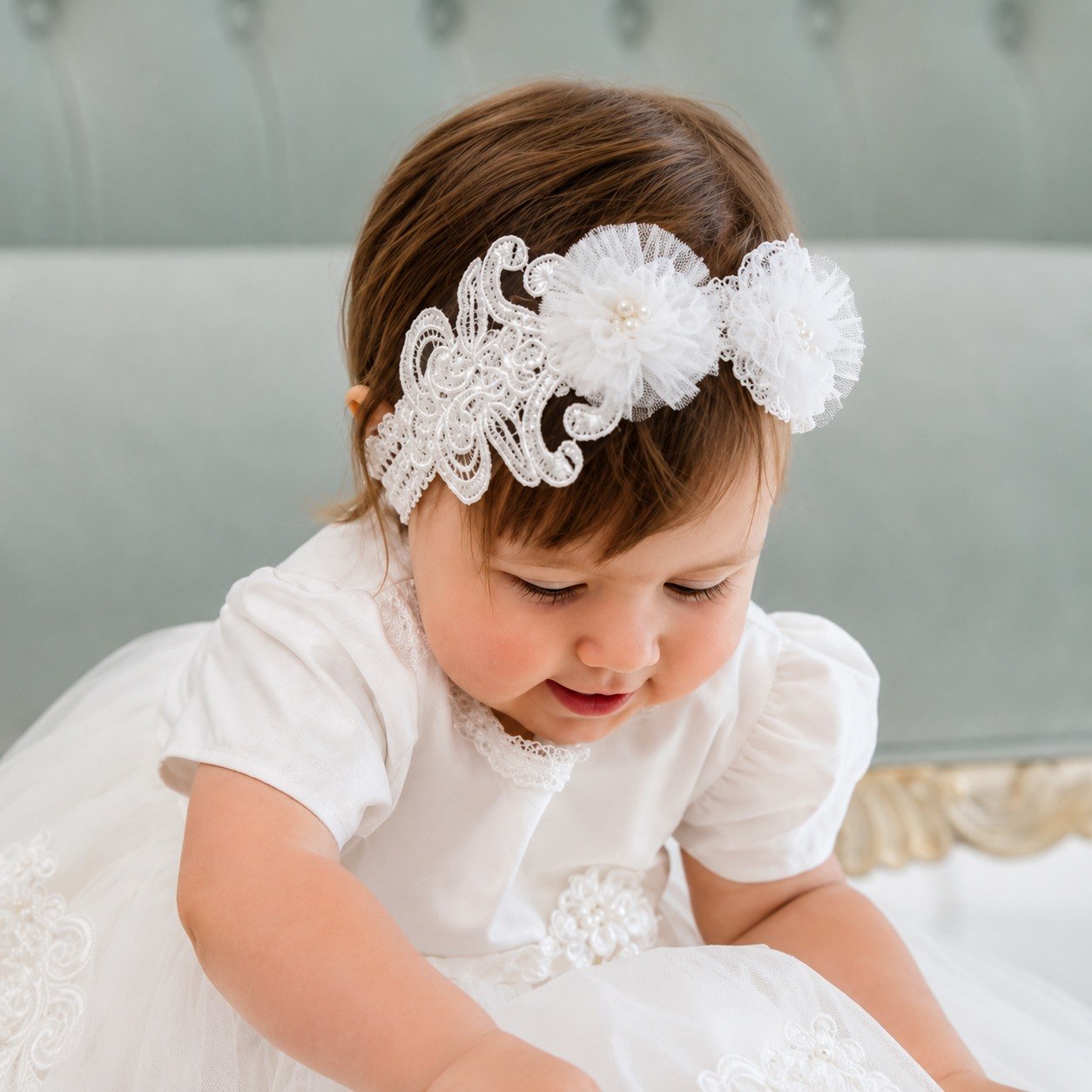 A toddler girl in a white dress wearing a white lace and fabric flower headband, looking down while sitting in front of a gray tufted headboard.