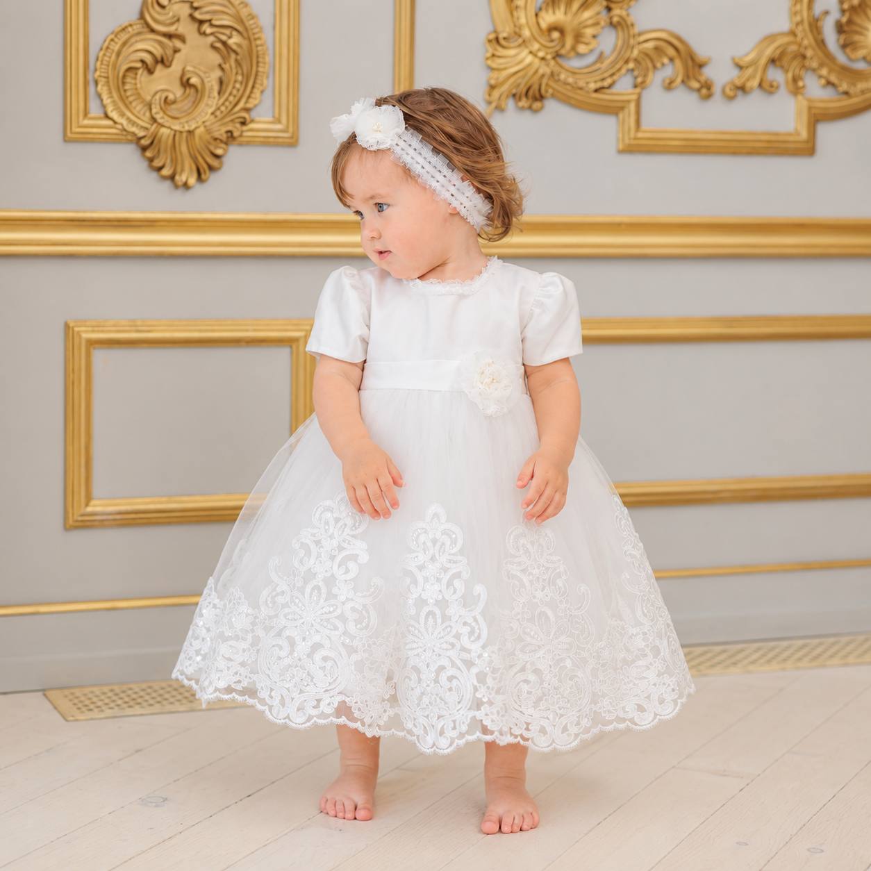 A toddler girl wearing a white first baptism dress with lace detailing and a matching white floral headband, standing in a room with gold-trimmed walls.