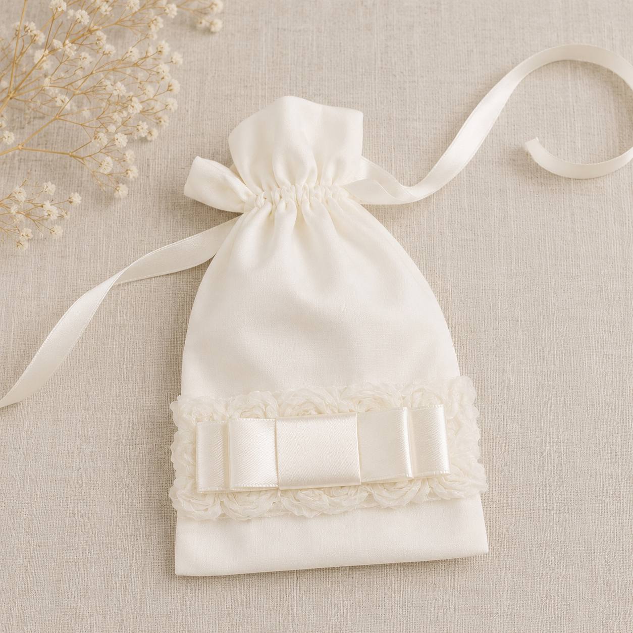An ivory satin drawstring pouch with a lace trim and ribbon bow, displayed on a textured beige linen background with dried baby's breath flowers.