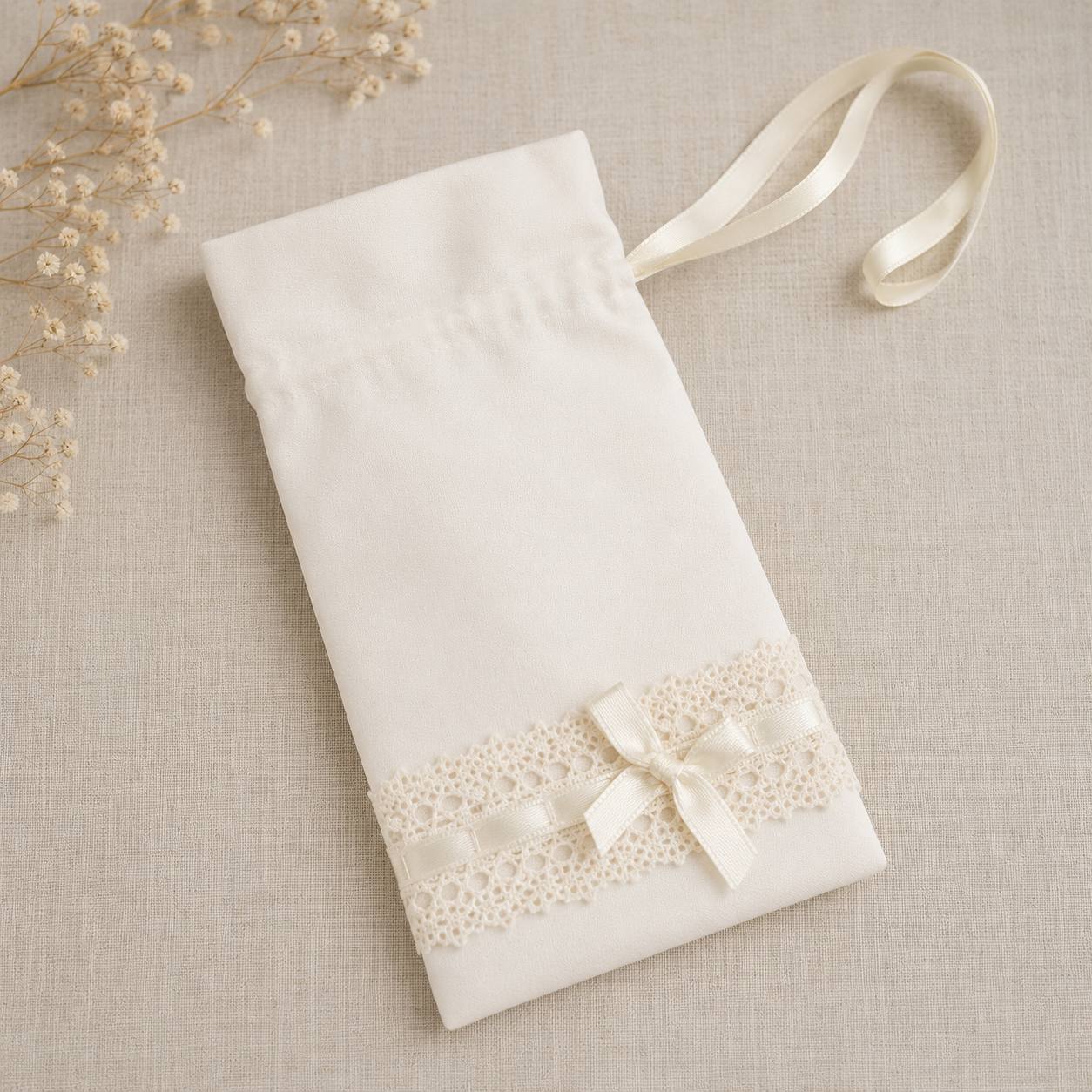 A cream-colored fabric drawstring pouch with a lace band and ribbon bow, displayed on a textured beige fabric background with dried baby's breath flowers.