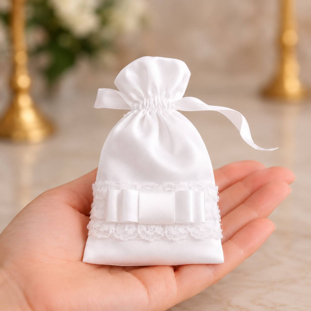 A small white satin drawstring pouch with lace trim and a ribbon bow, held in a person's hand against a softly blurred beige background.