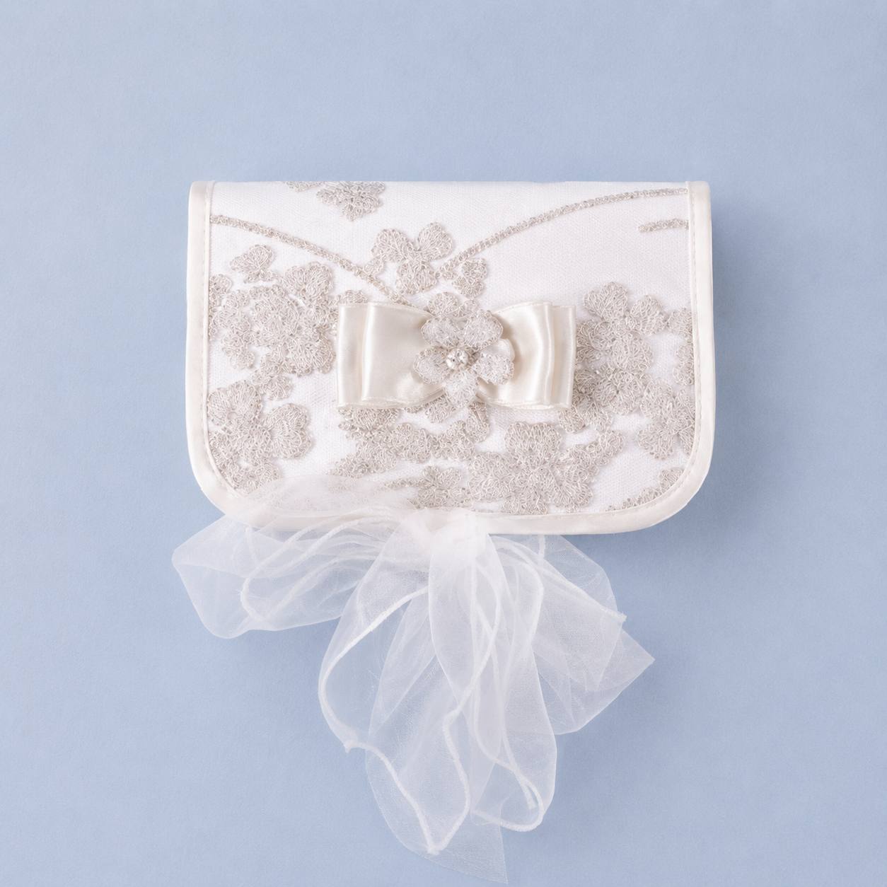 An ivory satin clutch purse with intricate floral lace embroidery, a central satin bow, and a long tulle ribbon tail, set against a solid light blue background.