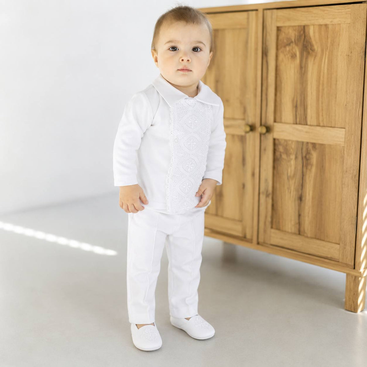 A toddler standing in a white traditional outfit with embroidered details on the chest, paired with white pants and white shoes, in front of a wooden cabinet.