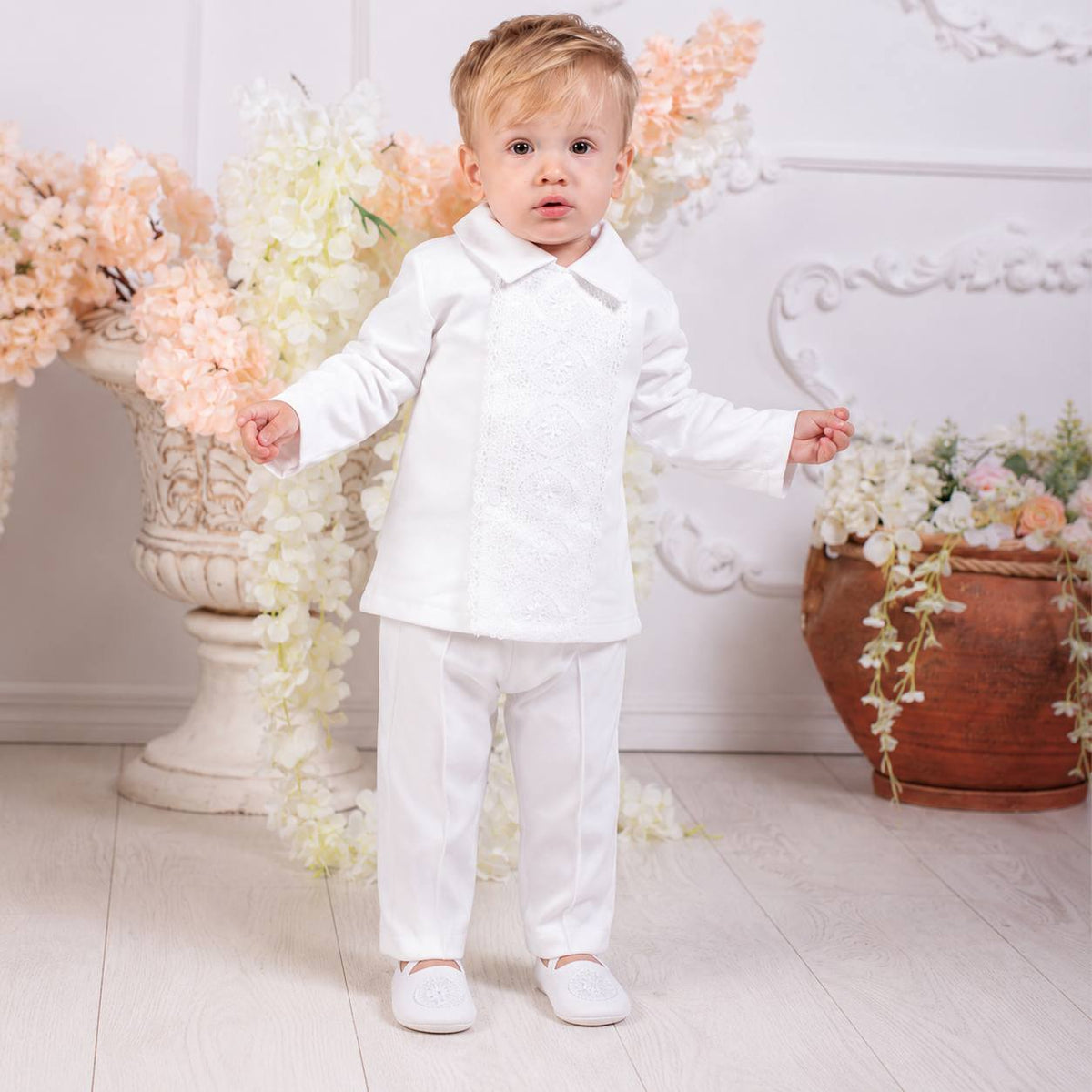 A toddler boy wearing a white formal outfit consisting of a long-sleeve collared shirt and matching trousers, standing in a decorated room with floral arrangements.