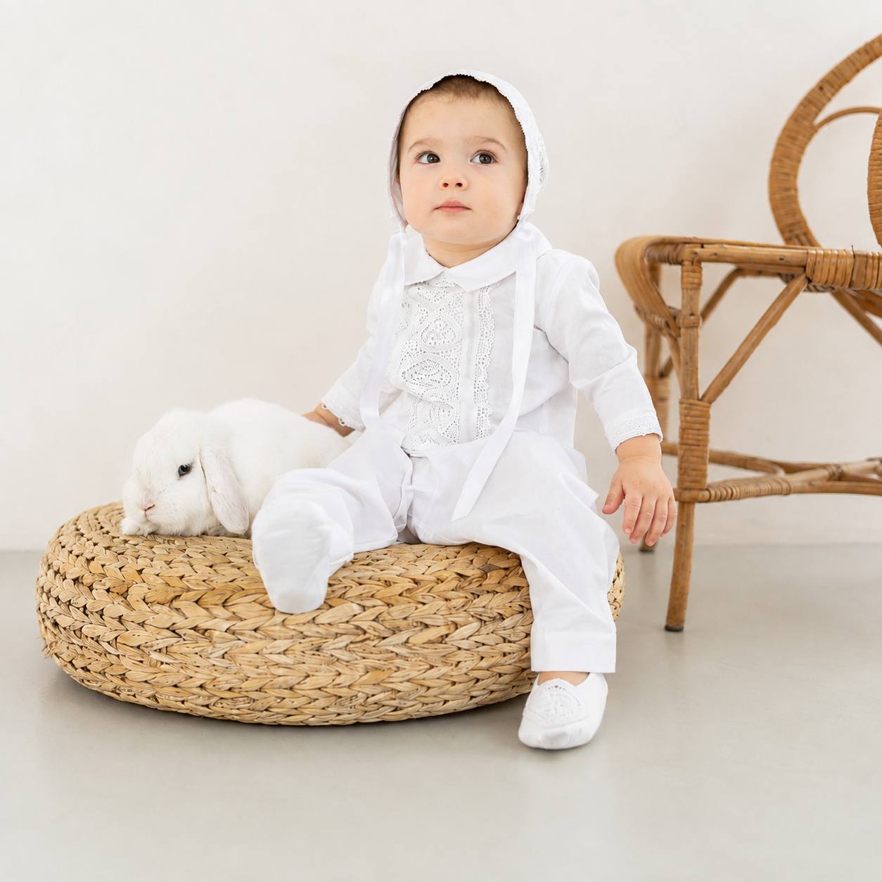 A baby wearing a white traditional outfit with a bonnet and white shoes sits on a woven straw pouf next to a white rabbit, with a wicker chair in the background.