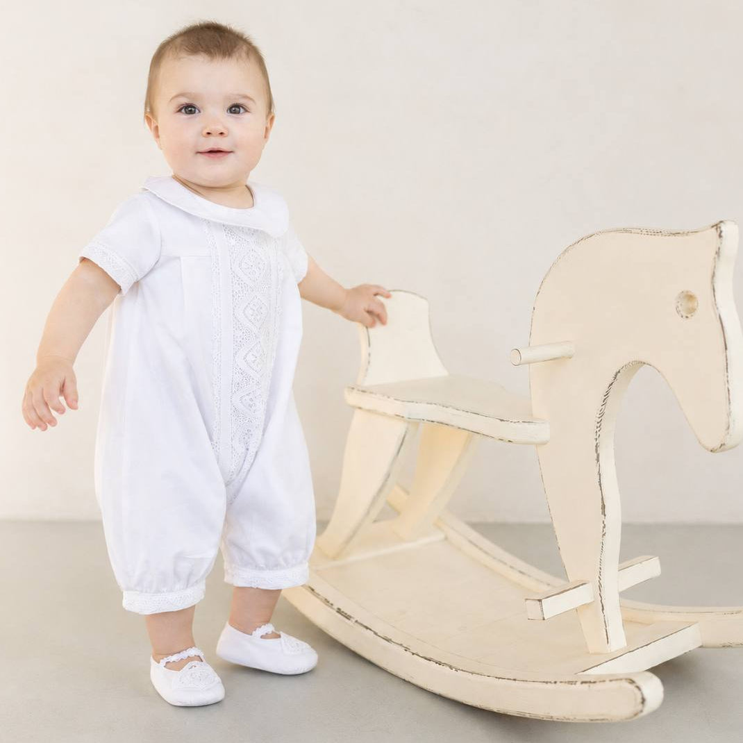 A baby wearing a white embroidered romper and white shoes stands next to a distressed cream wooden rocking horse against a neutral studio background.