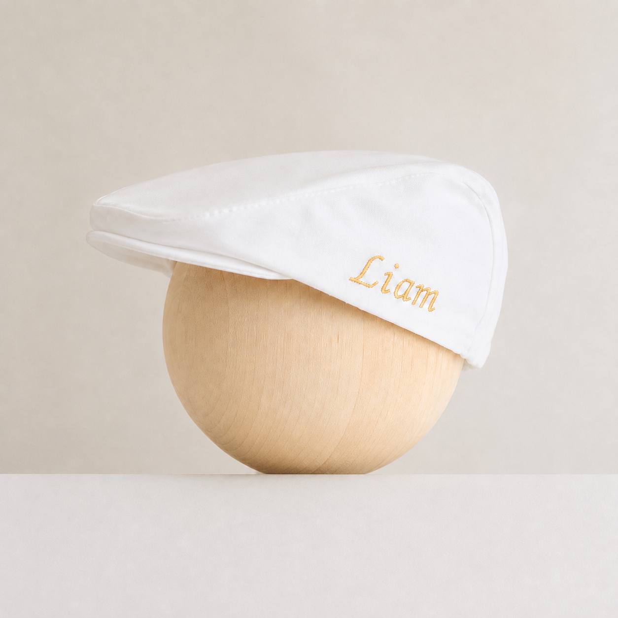 A white flat cap with the name 'Liam' embroidered in gold on the side, displayed on a light wood mannequin head against a neutral beige background.