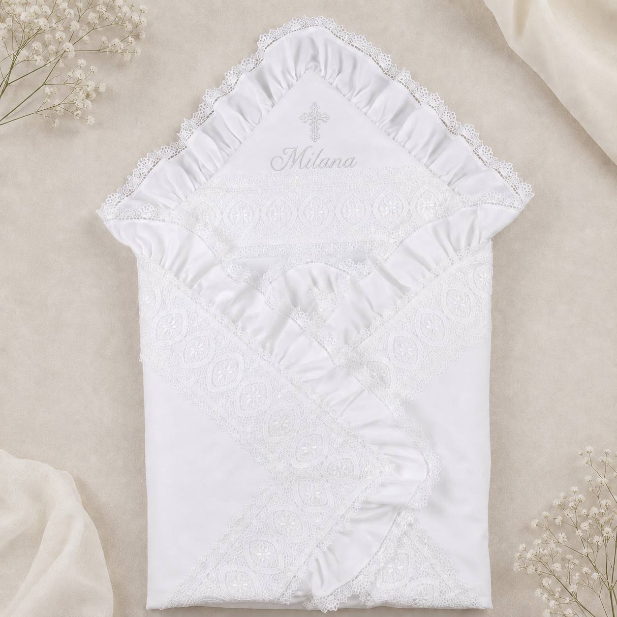 A white baby baptismal blanket with lace trim and embroidered details, laid flat on a beige fabric background with small white flowers.