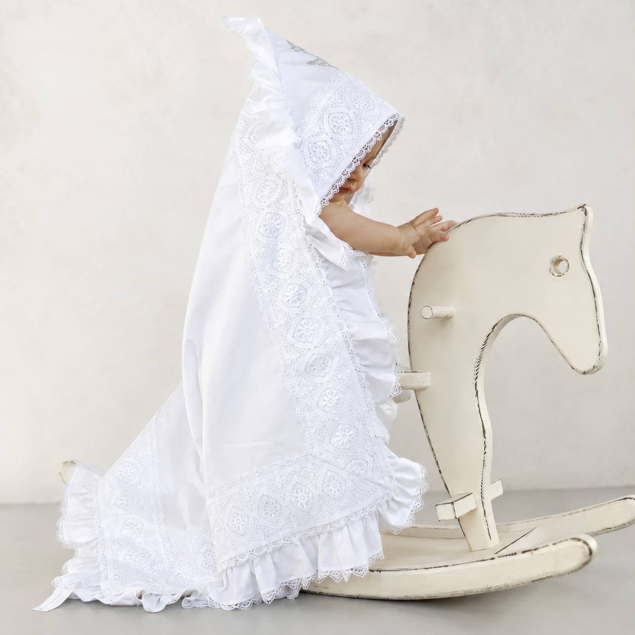 A baby wearing a white satin and lace baptism blanket, sitting on a distressed cream-colored wooden rocking horse against a neutral beige background.